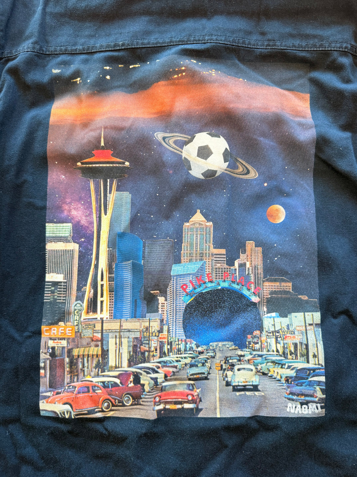 A close-up of a dark navy blue jacket featuring a large, colorful graphic print on the front. The graphic depicts a retro-futuristic Seattle scene with the Space Needle, a soccer ball planet, a ringed planet, and classic cars on a highway under a starry sky.