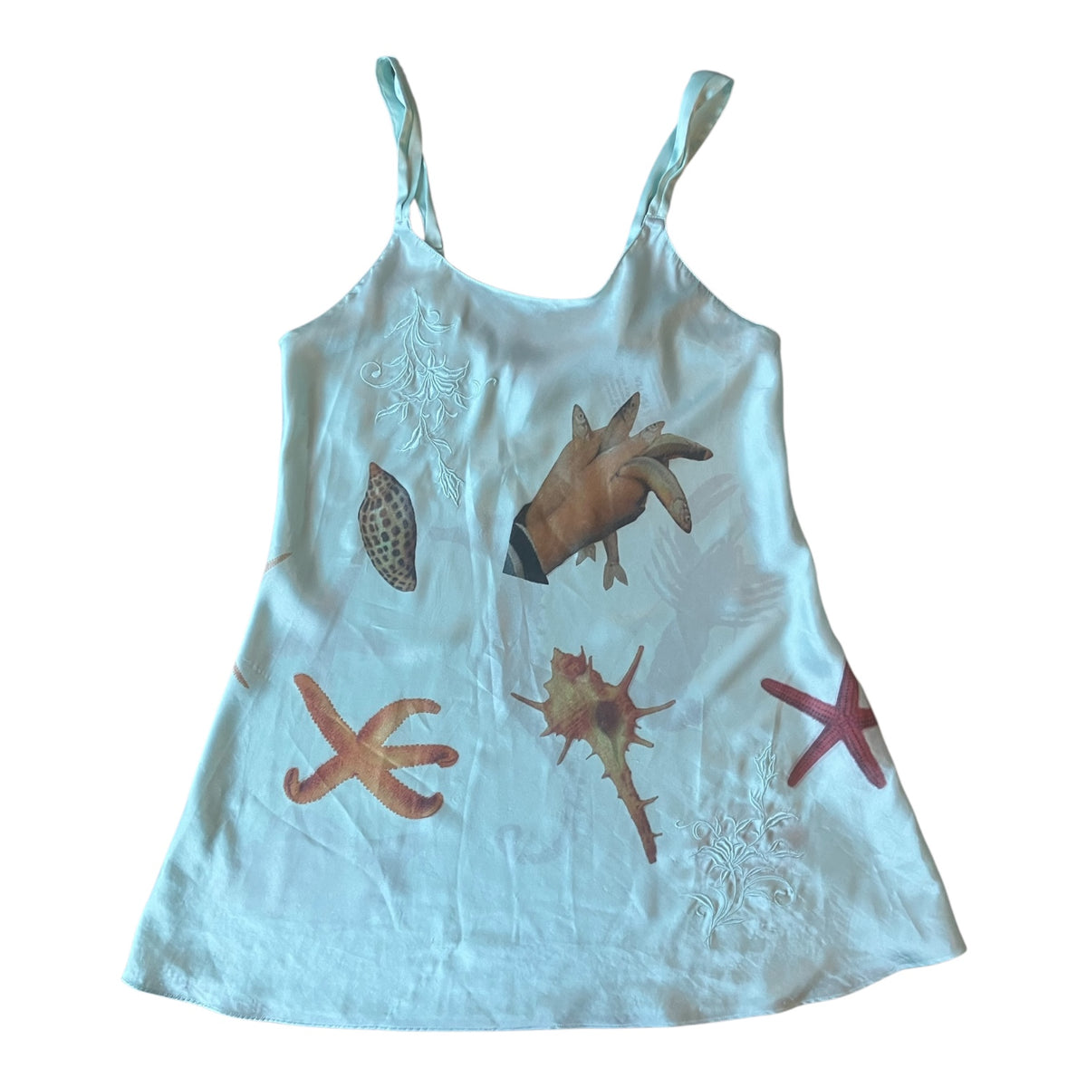 A light blue sleeveless short slip dress with a beach print featuring seashells, starfish, and a conch shell, laid flat against a white background.