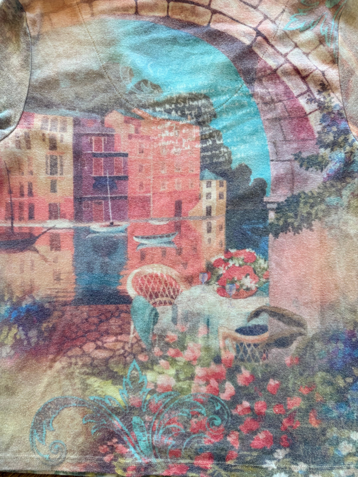 A close-up of a printed textile featuring a scenic coastal town with an archway, blue water, pink and red buildings, flowers, and outdoor seating. The fabric has a mottled, vintage-toned texture.