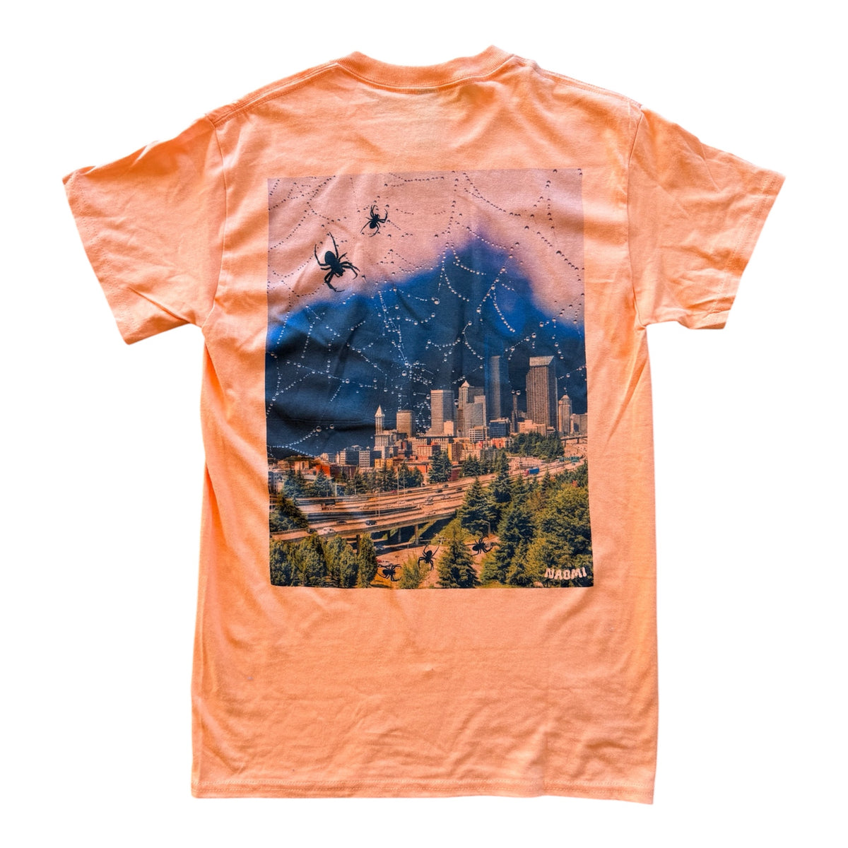An orange crewneck t-shirt shown flat against a white background, featuring a large rectangular graphic print on the back depicting a spiderweb over a city skyline at dusk.