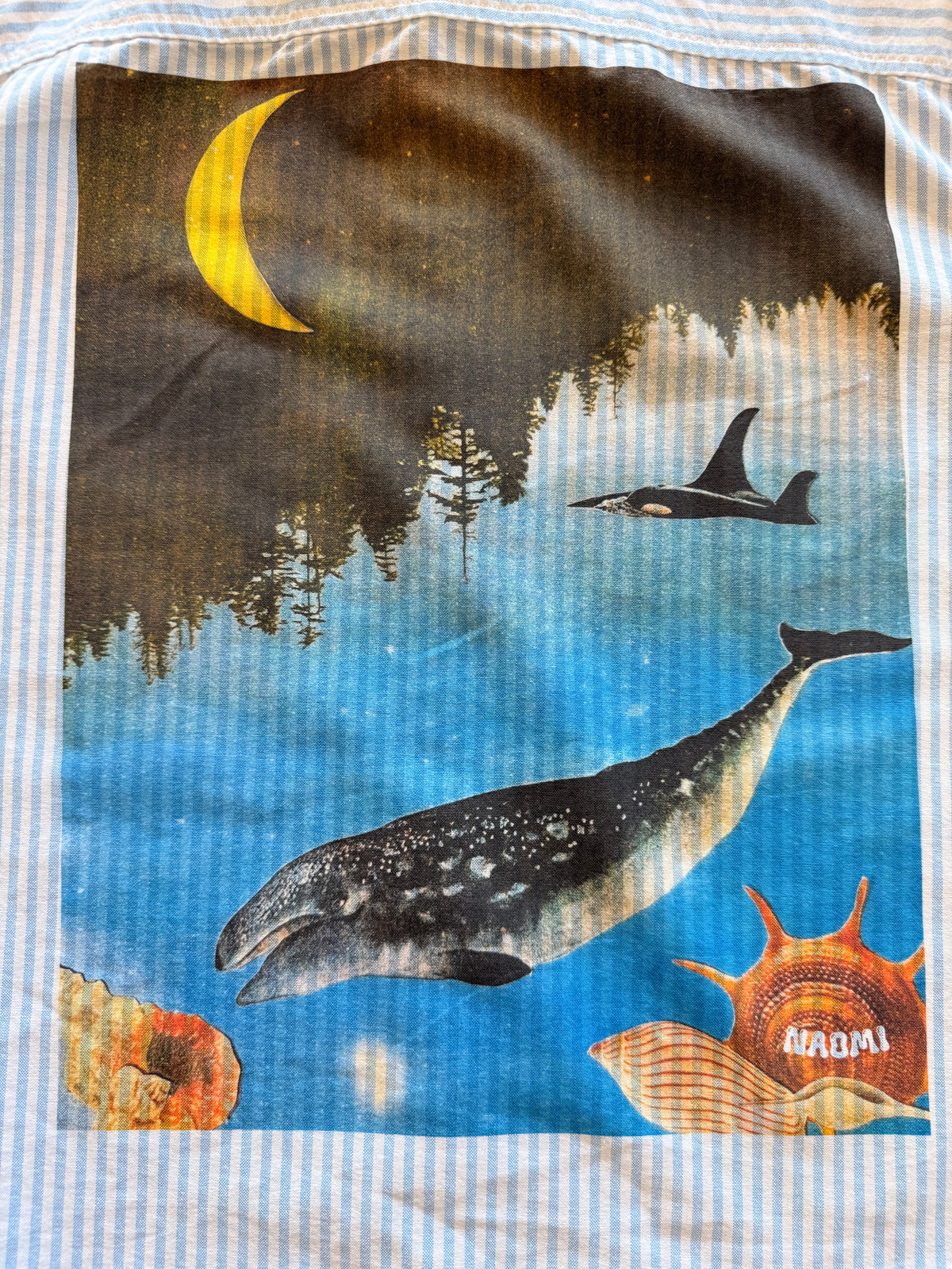 A close-up of a square fabric print featuring a whale swimming in blue water under a crescent moon, with a shark, trees, and shells. The print is on a white and light blue striped textile.