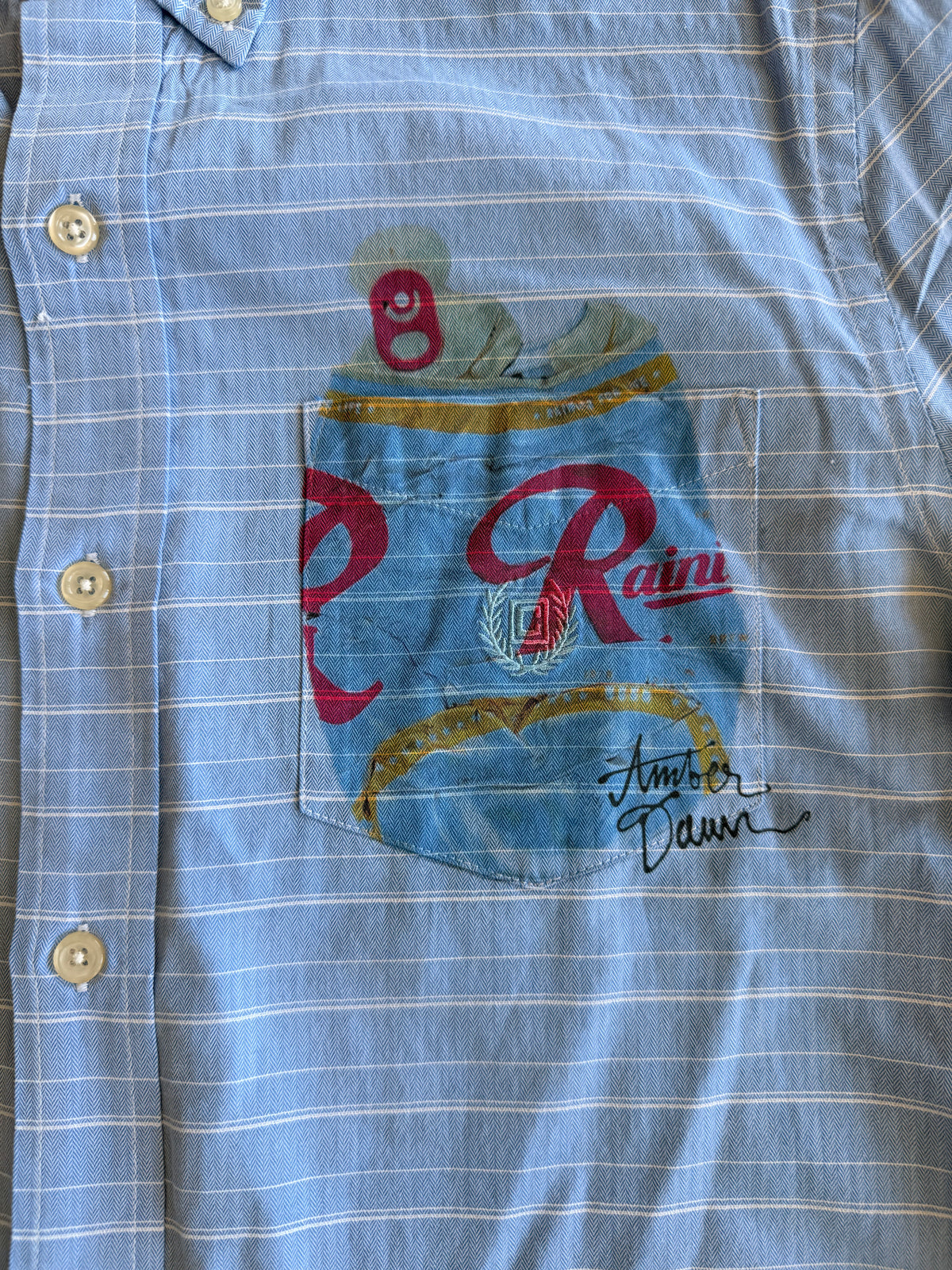 Close-up of a light blue and white striped button-down shirt featuring a printed Rainier brand beer can pocket design with red lettering and a small can graphic 