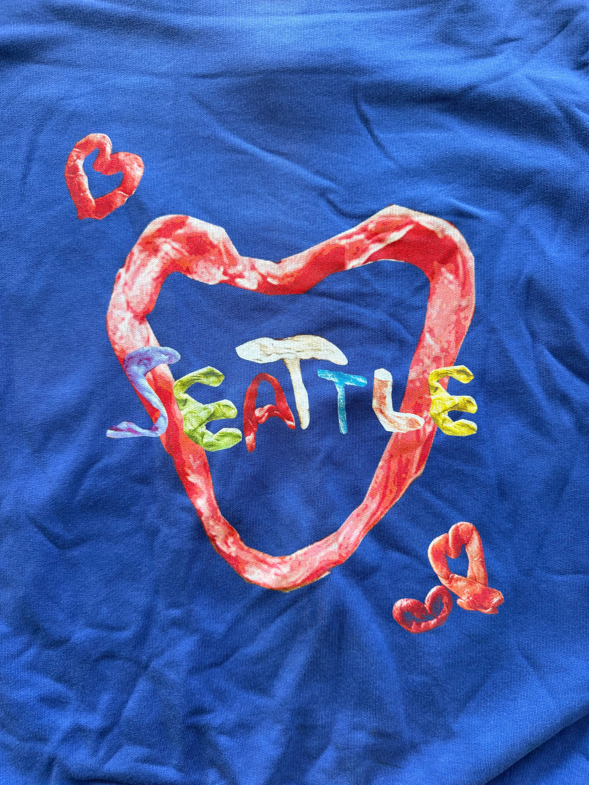 A close-up of a blue fabric surface featuring a hand-painted red heart outline with the word 'SEATTLE' written inside in multicolored letters, along with small red heart motifs in the corners.