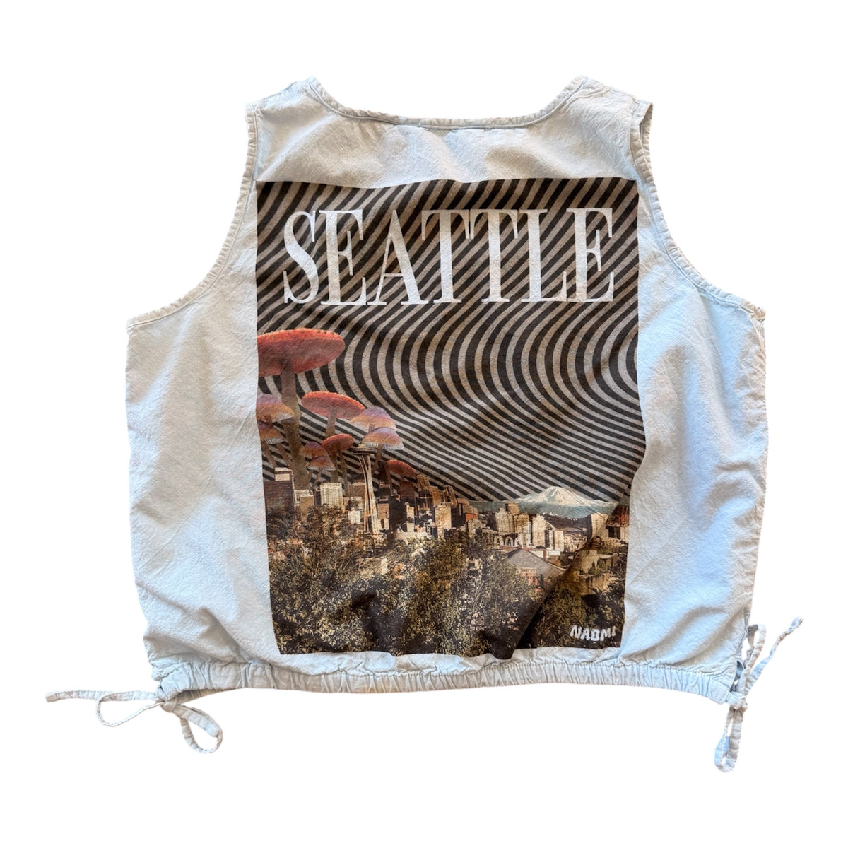 A sleeveless cropped vest in a light beige linen-like fabric, featuring a large graphic print on the front with the word 'SEATTLE' in white text over a black-and-white striped awning and a cityscape with red mushrooms. The vest has drawstring ties at the sides and is photographed flat against a white background.