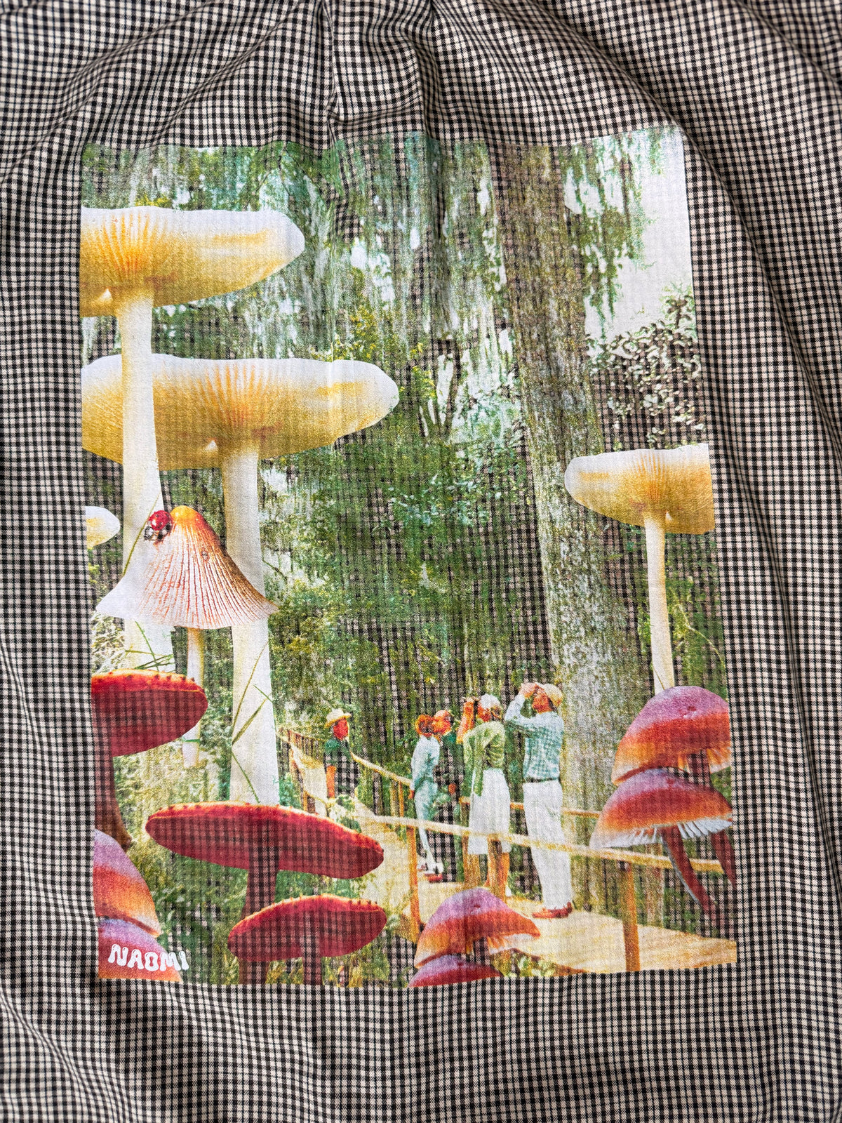 A black and white gingham patterned shirt featuring a large rectangular front patch print of a whimsical forest scene with giant mushrooms, moss trees, and people on a wooden walkway.