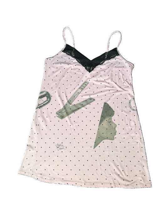 A light pink camisole nightshirt with black polka dots, a black lace V-neckline, and gray graphic illustrations of nail clippers, a safety pin and a profile silhouette.