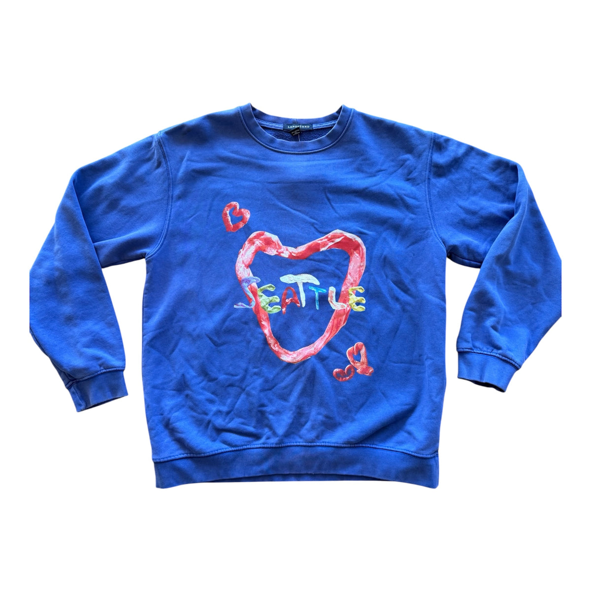 A blue crewneck sweatshirt featuring a graphic of a red heart outline with the word 'SEATTLE' written inside in a multicolored, textured font, accented by small red hearts on the sleeves, shown flat on a white background.