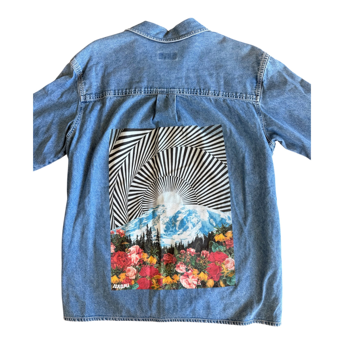 A light blue denim shirt jacket shown from the back, featuring a large graphic print of a mountain landscape with a sunburst and a bed of colorful flowers.