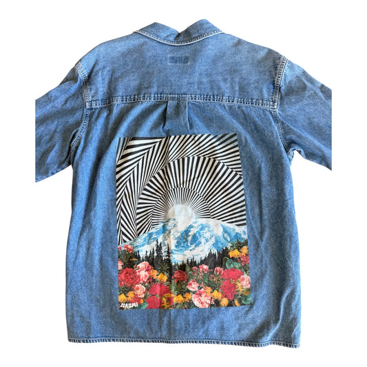 A light blue denim shirt jacket shown from the back, featuring a large graphic print of a mountain landscape with a sunburst and a bed of colorful flowers.