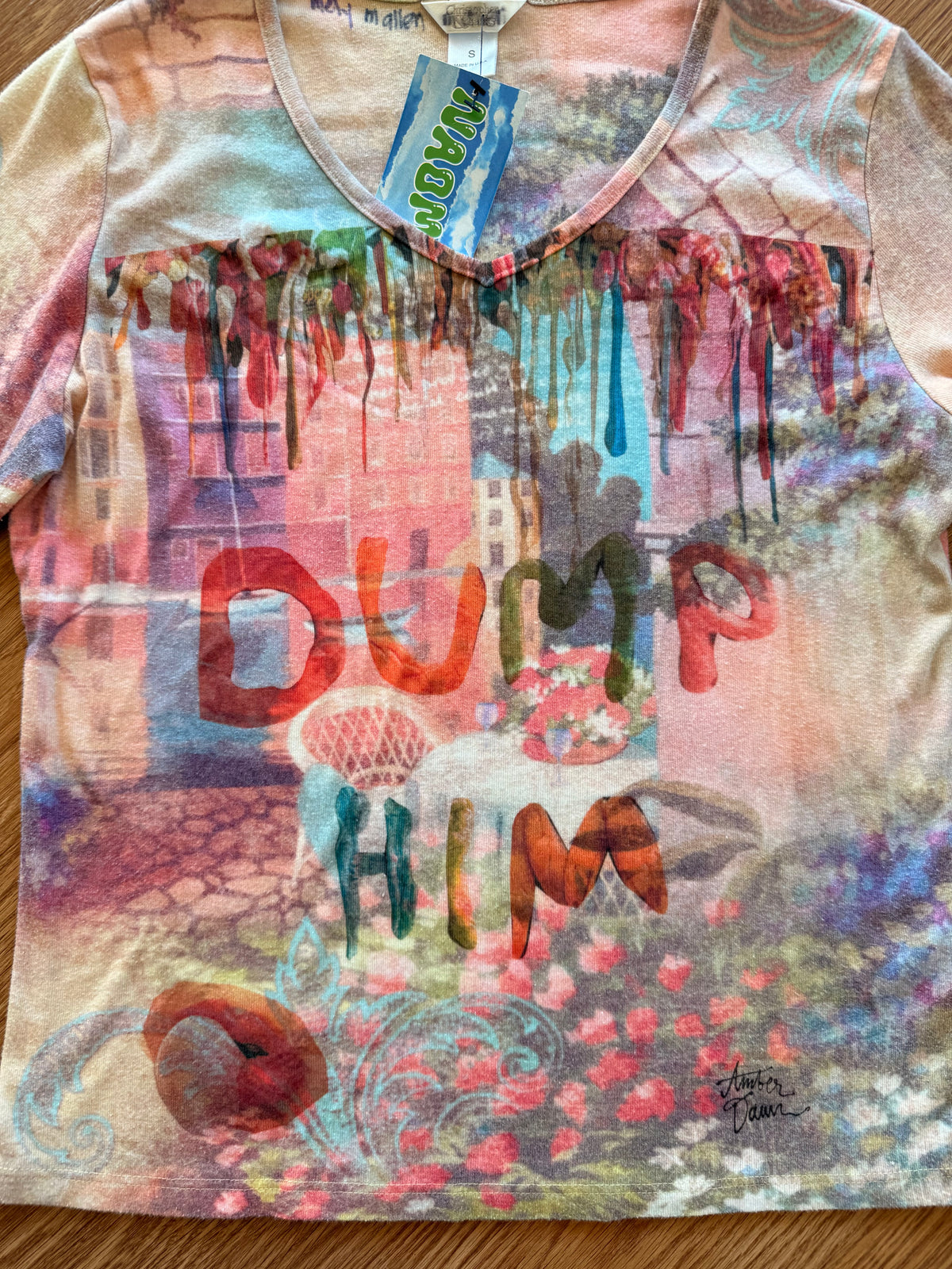 A short-sleeve V-neck graphic t-shirt featuring a colorful graffiti-style print with the words 'DUMP HIM' in large letters, flowers, and paint drips, laid flat on a wooden surface.