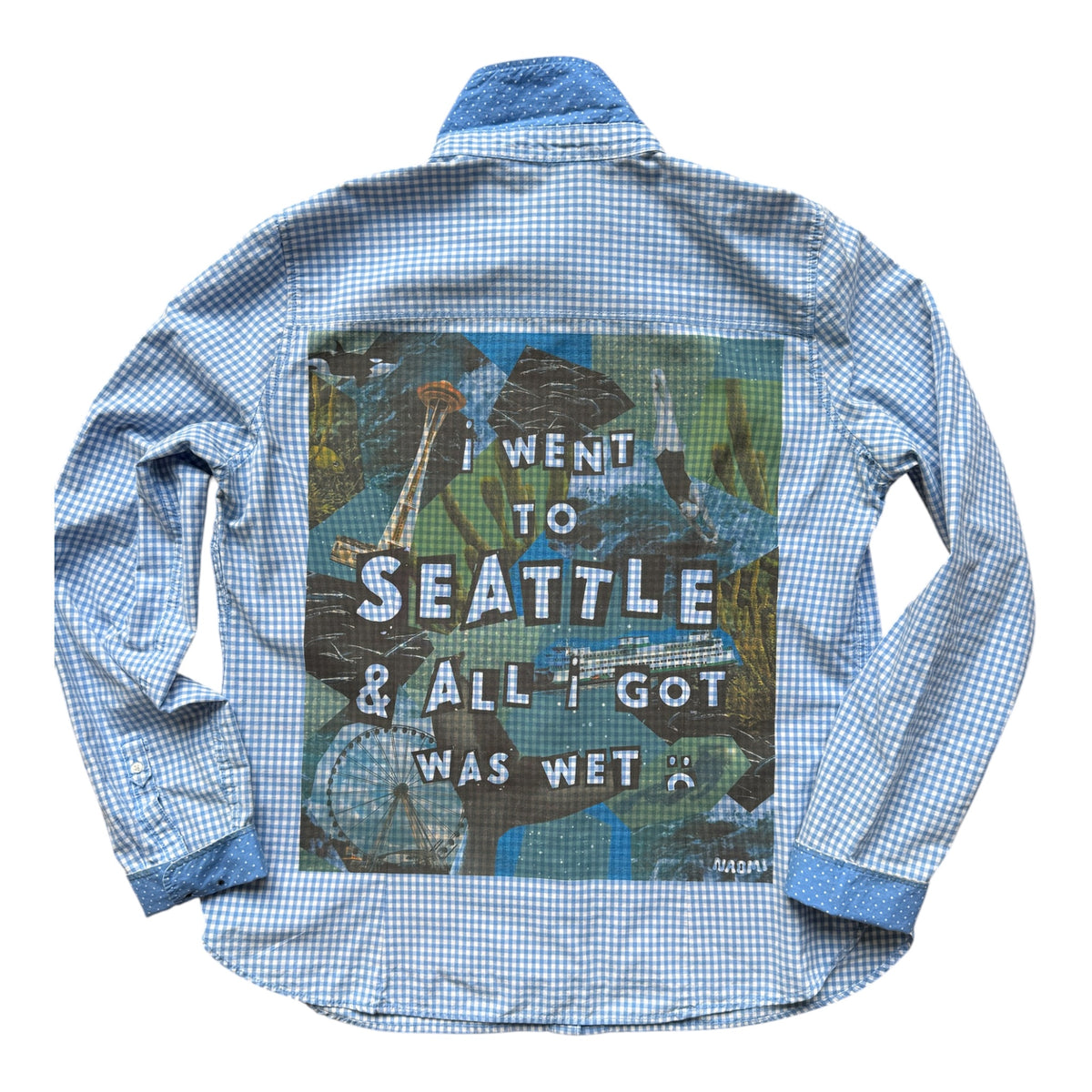 A light blue and white gingham long-sleeve button-up shirt shown flat against a white background, featuring a large graphic print on the back with a Seattle seascape and the text 'I WENT TO SEATTLE & ALL I GOT WAS WET'.