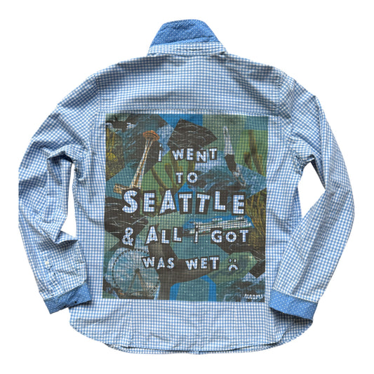 A light blue and white gingham long-sleeve button-up shirt shown flat against a white background, featuring a large graphic print on the back with a Seattle seascape and the text 'I WENT TO SEATTLE & ALL I GOT WAS WET'.