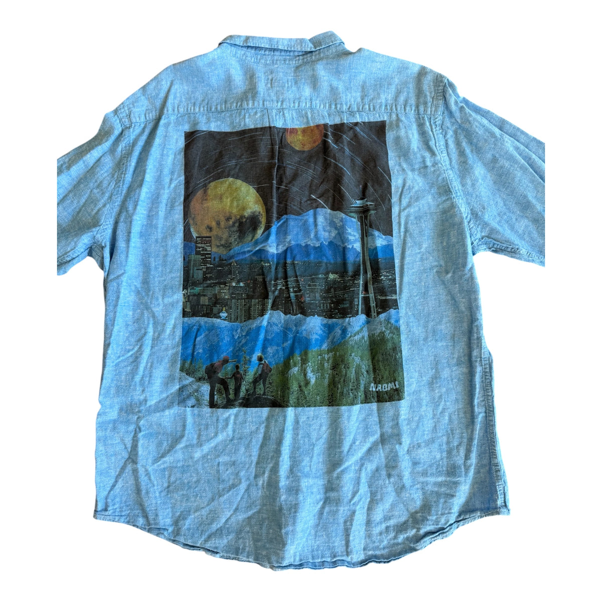 A light blue chambray short-sleeve button-up shirt shown from the back, featuring a large rectangular graphic print of a surreal landscape with mountains, a city skyline, and large yellow moons.
