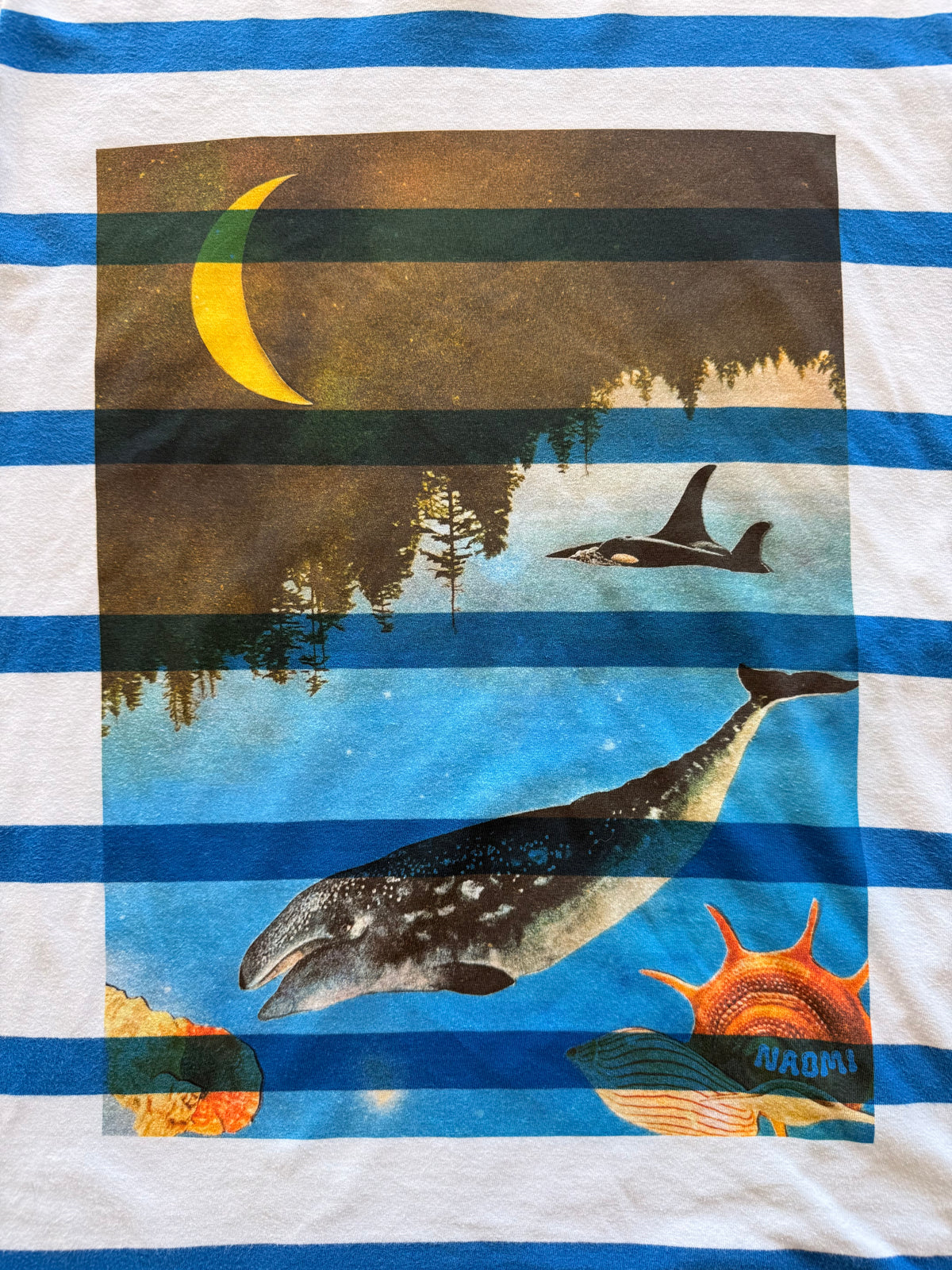 A close-up of a striped white and blue garment, likely a sweatshirt or hoodie, featuring a large rectangular graphic print. The print shows a whale in blue water, an orca, a crescent moon, trees, and a sun.