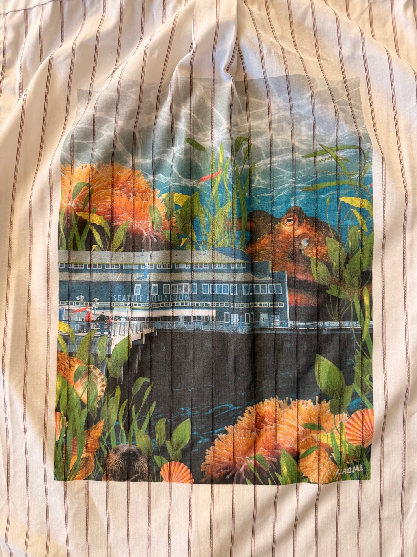 A square fabric print or patch featuring an underwater scene with the seattle aquarium, orange sea plants, and a large fish, placed on a cream-colored pinstriped fabric background.