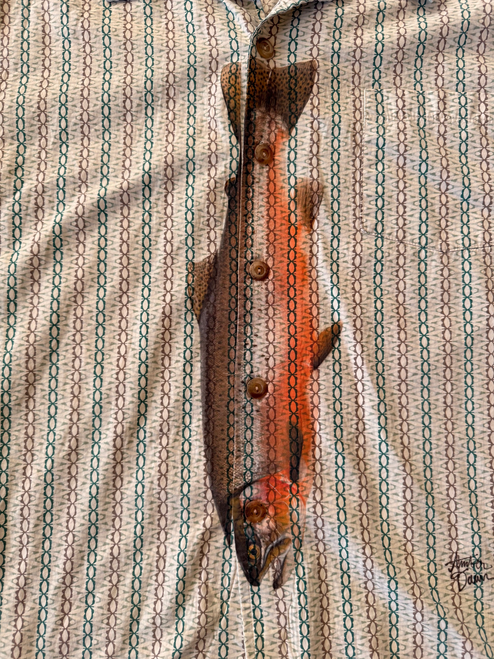 A whole rainbow trout lies vertically across a patterned button-up shirt. The shirt has a beige base with repeating vertical teal and dark green chain-link motifs.
