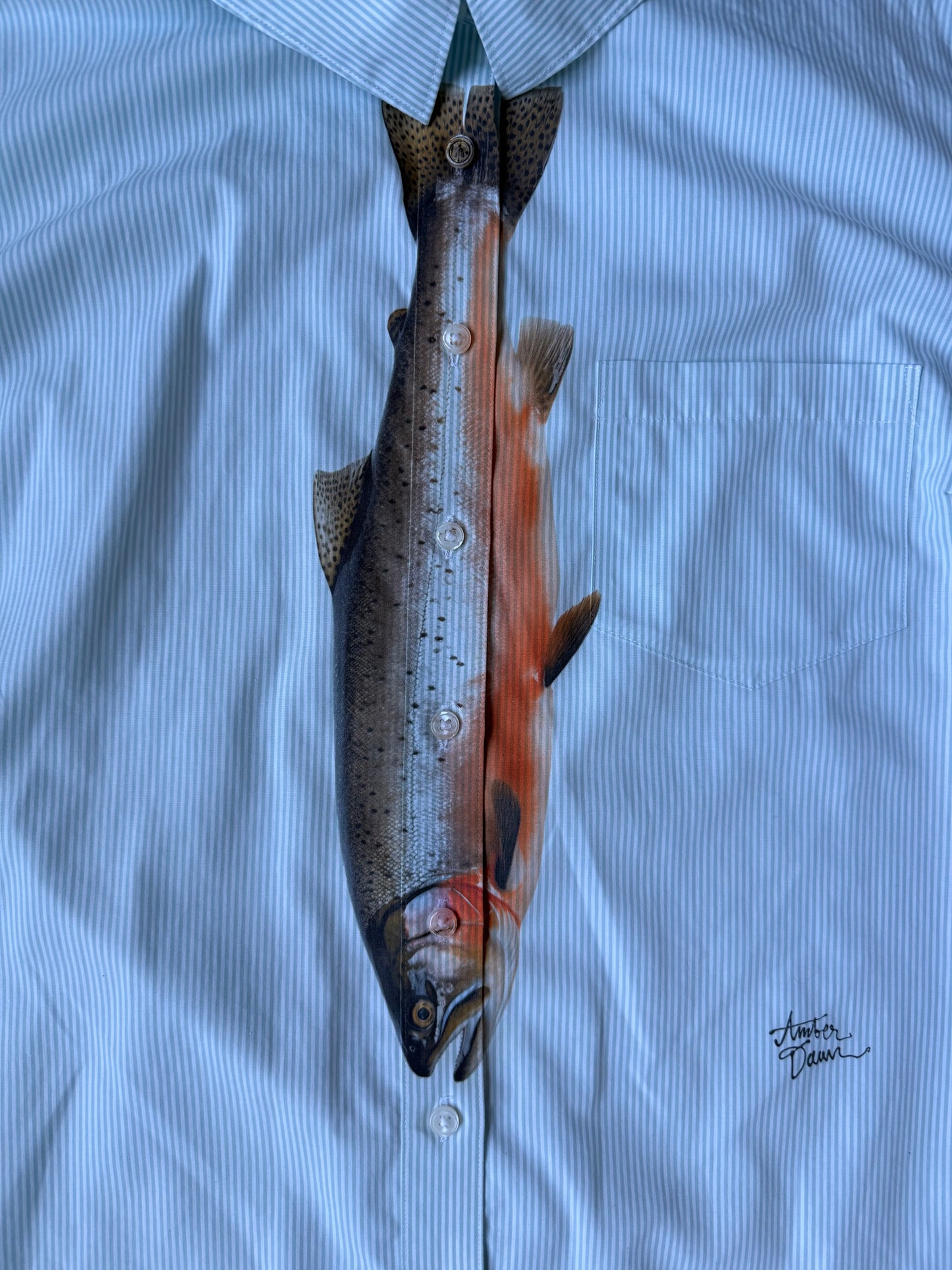 Up close of a light blue and white striped button down with a fish printed on the front to look like a tie 