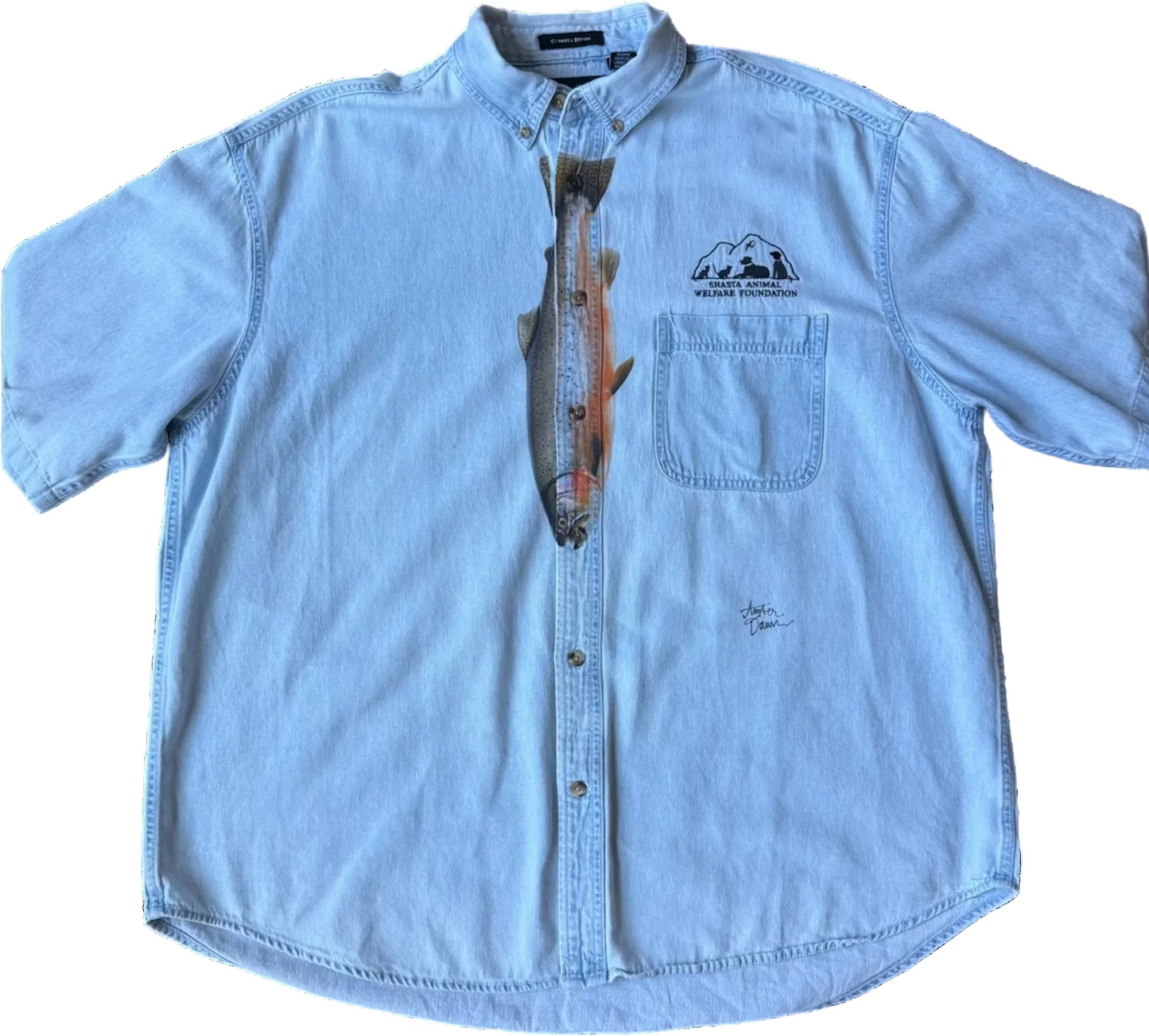 A light blue denim short-sleeve button-up shirt with a chest pocket and a printed fish graphic running vertically down the center front. The shirt features a ‘Shasta Animal Welfare Foundation' logo on the left chest.