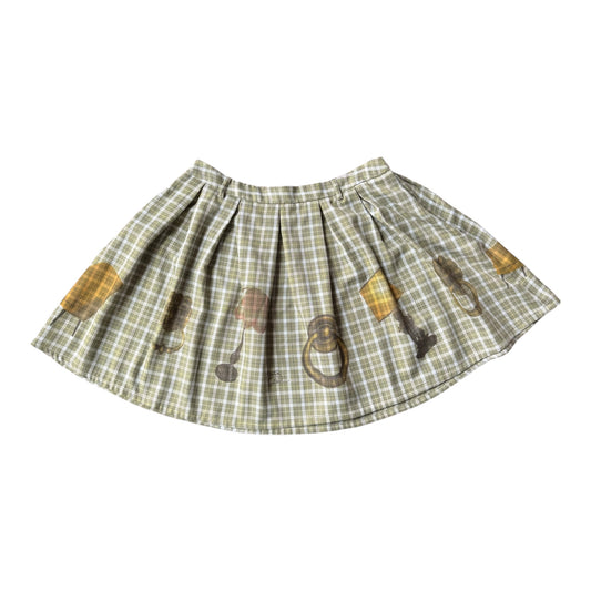 A pleated skirt in a muted green and beige plaid pattern, decorated with illustrations of vintage lamps and door knockers in yellow, brown, and gray, shown against a plain white background.