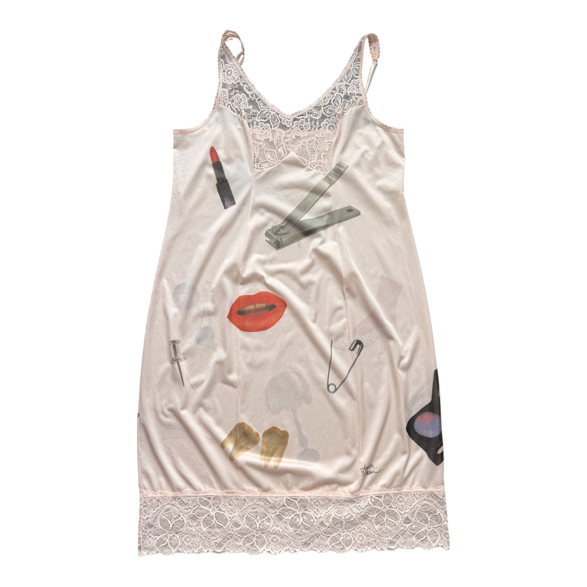 A light pink satin slip dress featuring a collage print of various makeup items, safety pins, and red lips, finished with a lace trim at the neckline and hem.