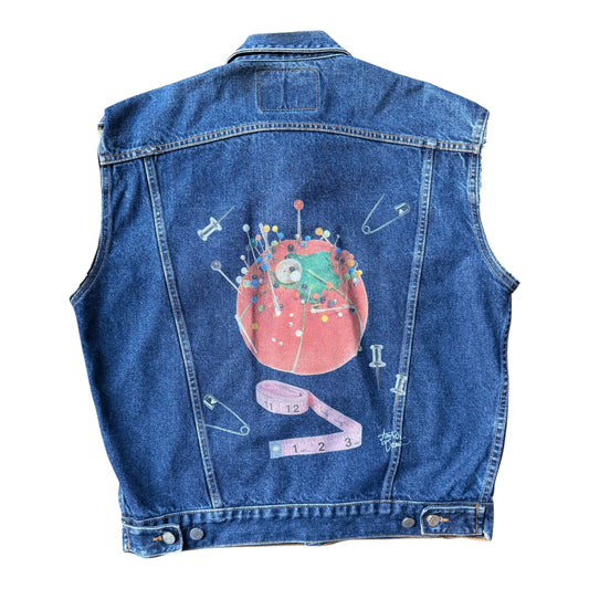 The back of a blue denim vest featuring a large graphic print of a sewing-themed pin cushion with a green character, safety pins, and a pink measuring tape on a white background.