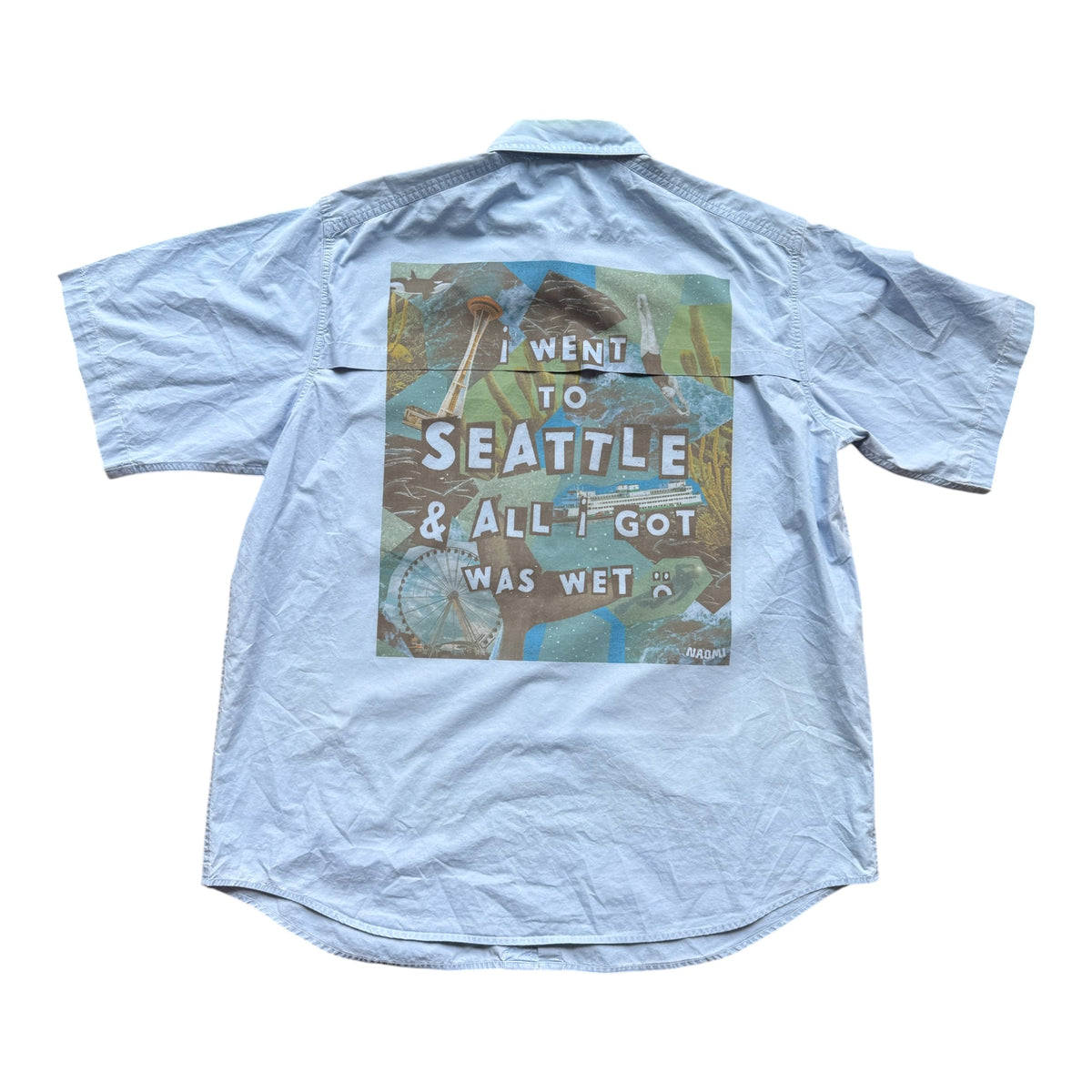 A light blue short-sleeve button-up shirt shown flat against a white background, featuring a large graphic print on the back with the text 'I WENT TO SEATTLE & ALL I GOT WAS WET' over a colorful Seattle-themed illustration.