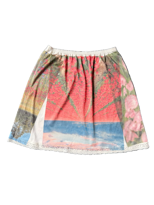 A short A-line skirt with a colorful scenic print featuring red trees, a blue sky, and pink flowers, finished with a white lace trim at the hem.