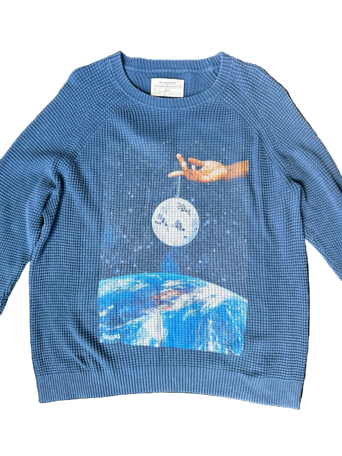 A blue waffle-knit crewneck sweatshirt featuring a graphic print of a hand holding a moon above a blue and white Earth, displayed flat against a white background.