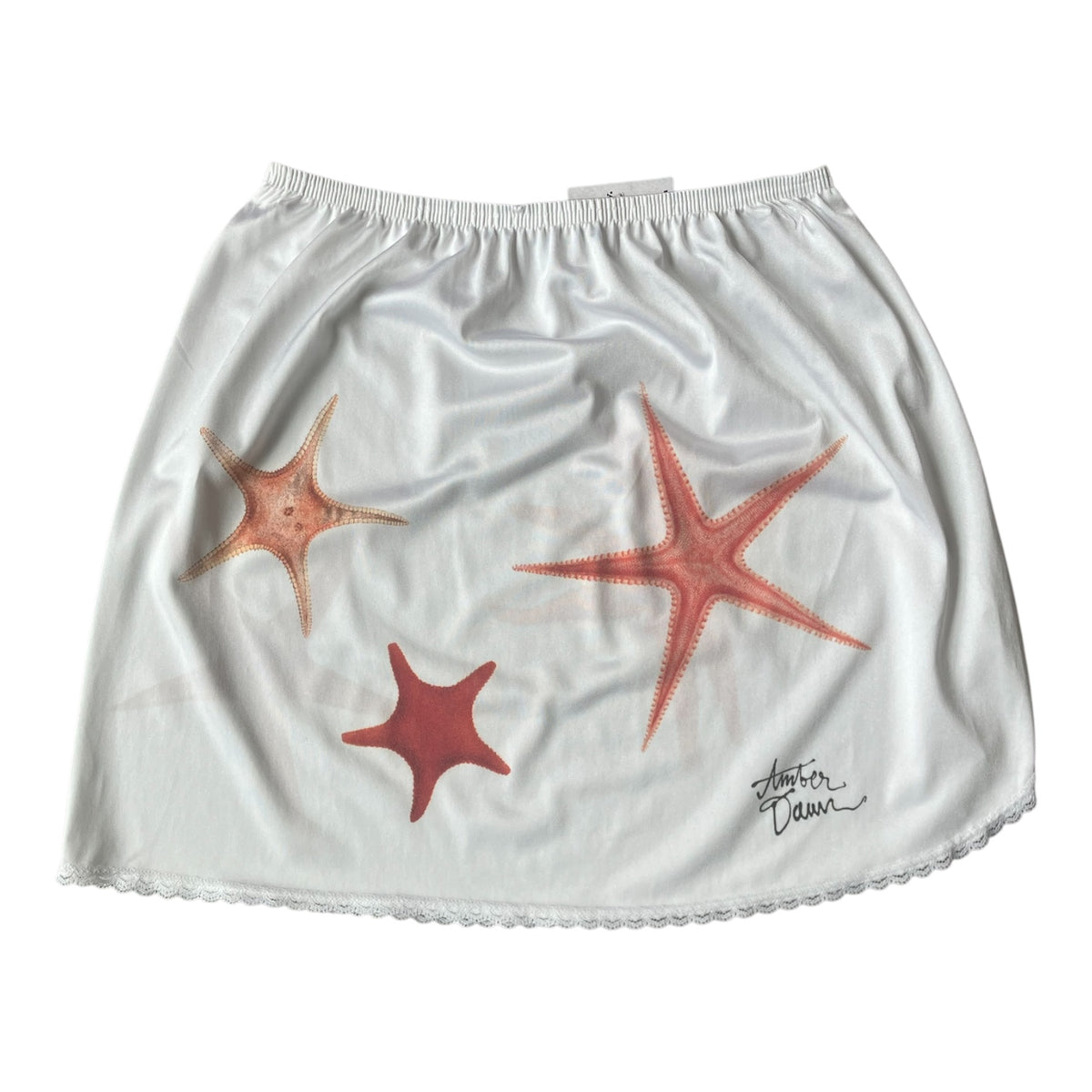 White satin-like skirt with a scalloped hem, featuring three hand-painted starfish in shades of orange, red, and pink, and a black script signature on the lower right.