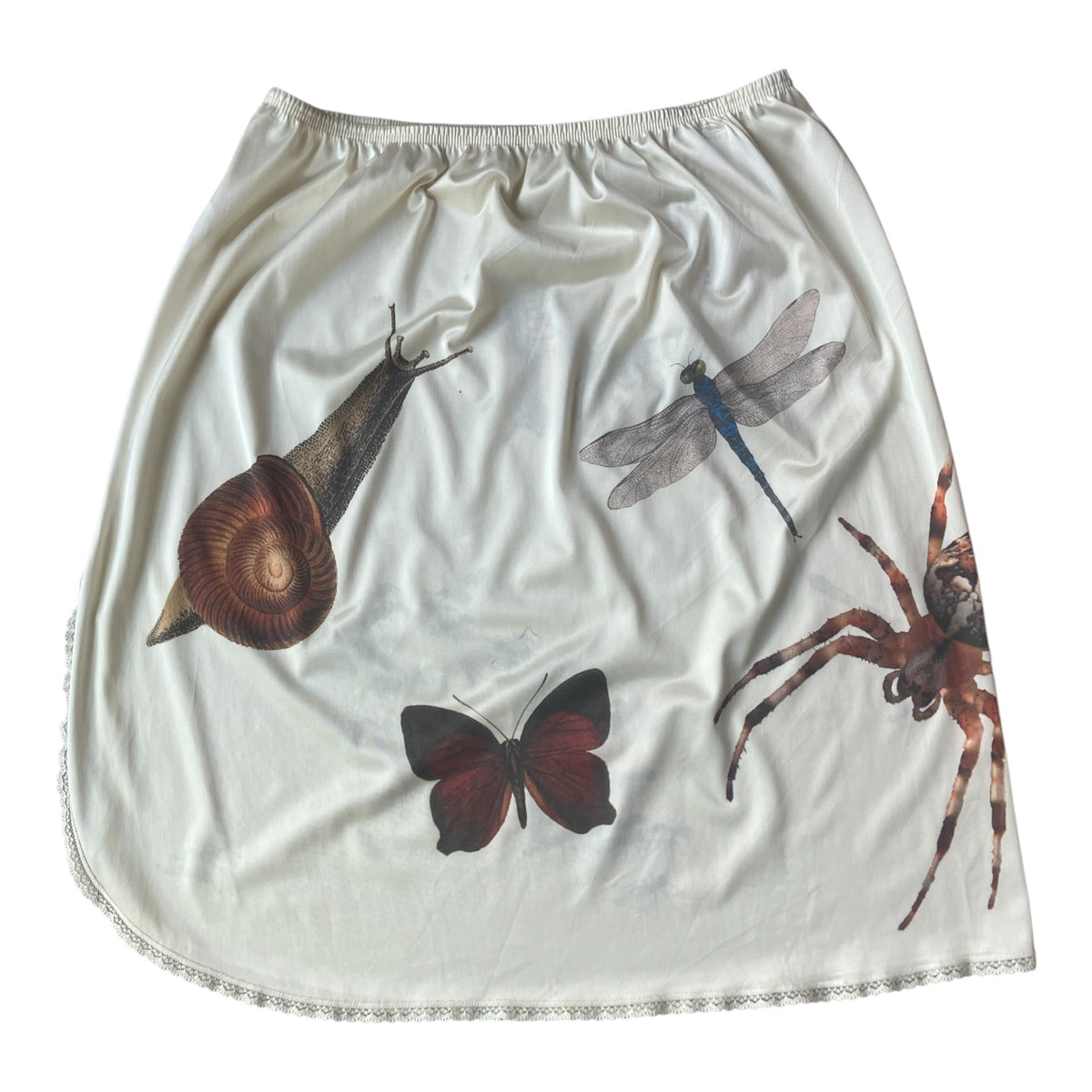 An off-white satin mini skirt featuring a printed design of various insects, including a snail, dragonfly, butterfly, and spider, with a ruffled trim along the hem.