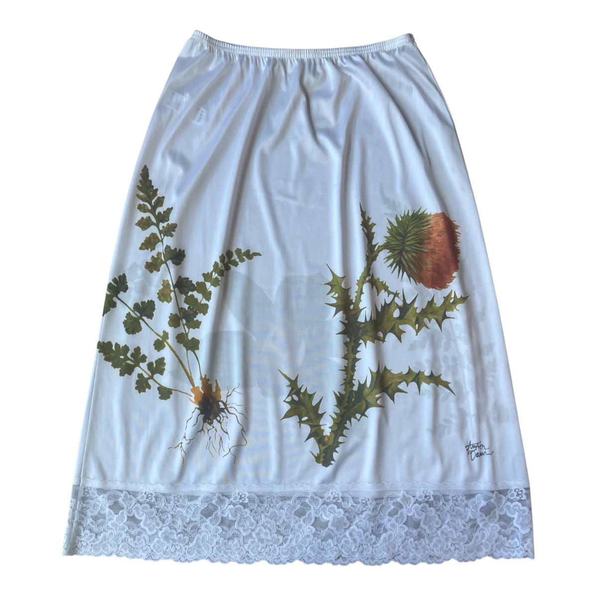 A white midi skirt featuring a botanical print of green thistles and ferns, finished with a white lace hem.