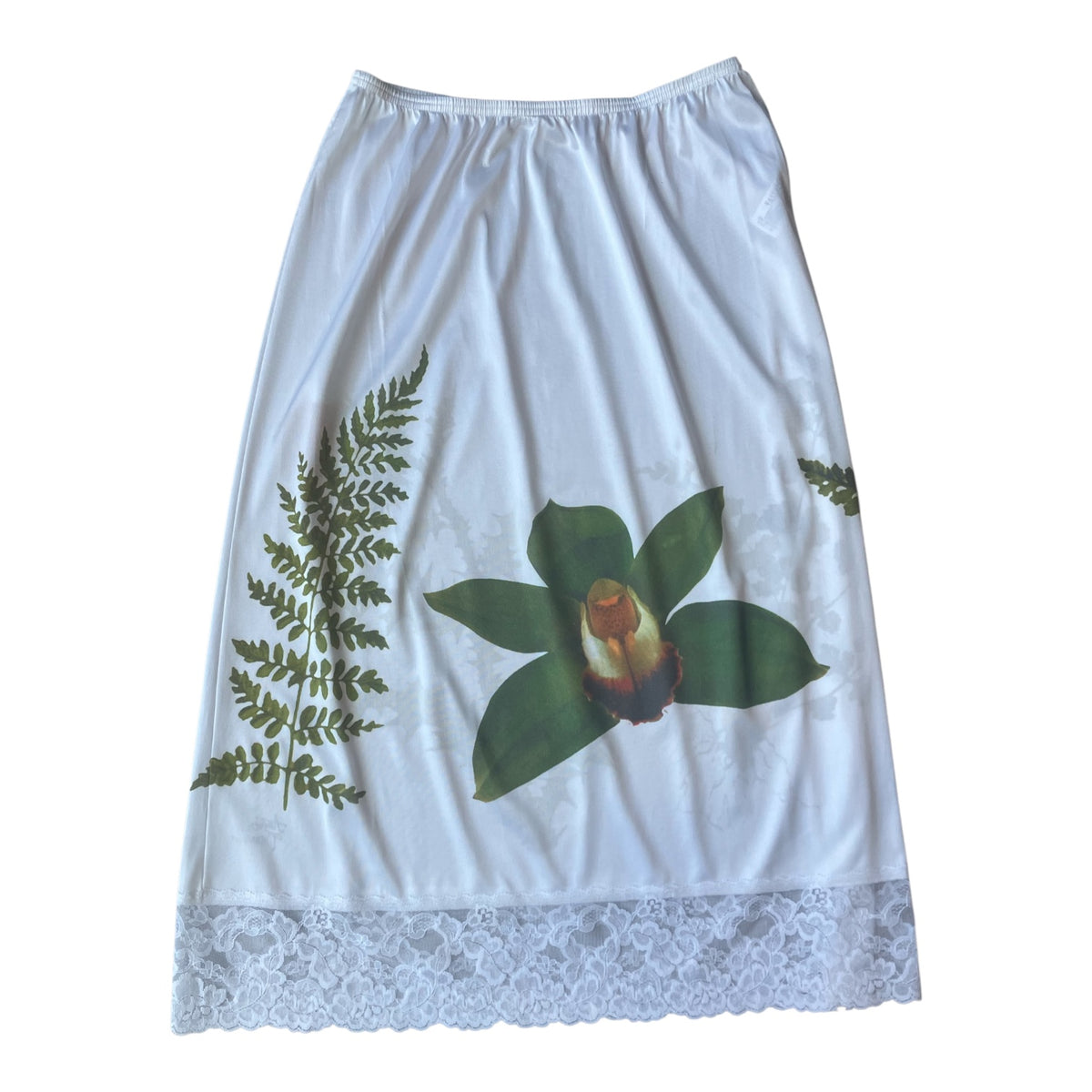 A white midi skirt featuring a green orchid and fern botanical print, finished with a white lace hem.