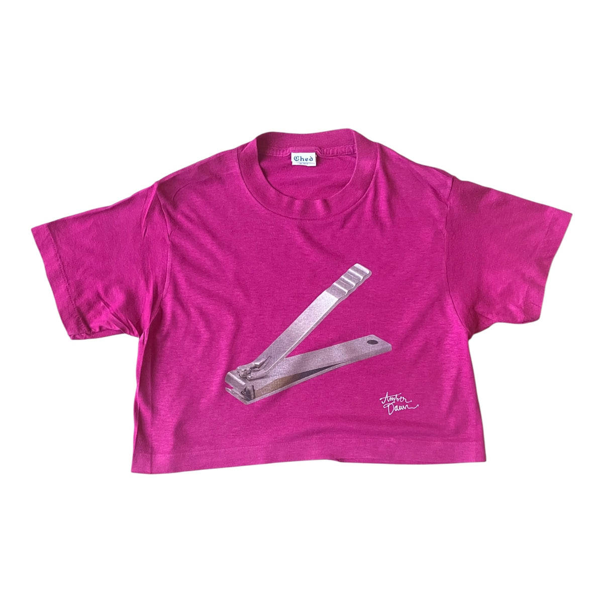 A bright pink short-sleeve crop top t-shirt featuring a graphic of silver nail clippers on the front, laid flat against a white background.