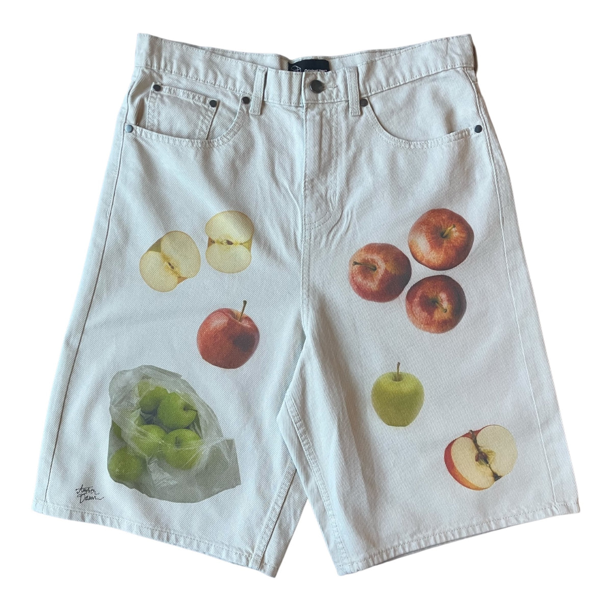 Off-white denim shorts featuring a printed pattern of various apples, including whole red apples, sliced apples showing pale yellow flesh, a green apple, and a clear plastic bag filled with green apples.