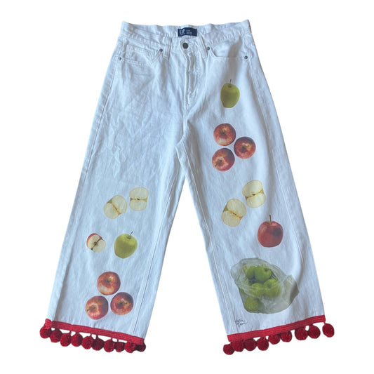 White denim cropped pants featuring a colorful print of apples and a bag of green apples, finished with red pom-pom trim at the hems.